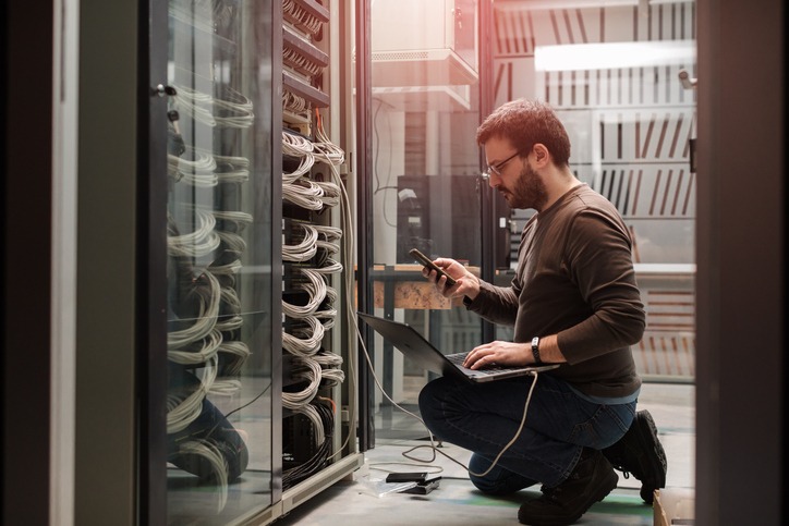 man kneeling in server room on phone with wire connected to his laptop